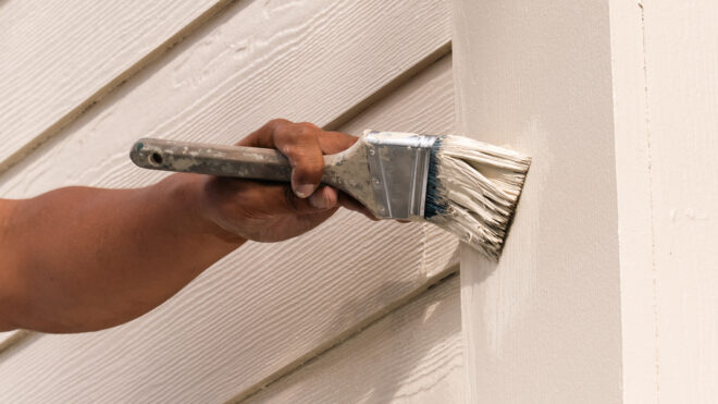 Hand holding a paintbrush applying a second coat of paint to cream-colored lap siding after the proper drying time between coats