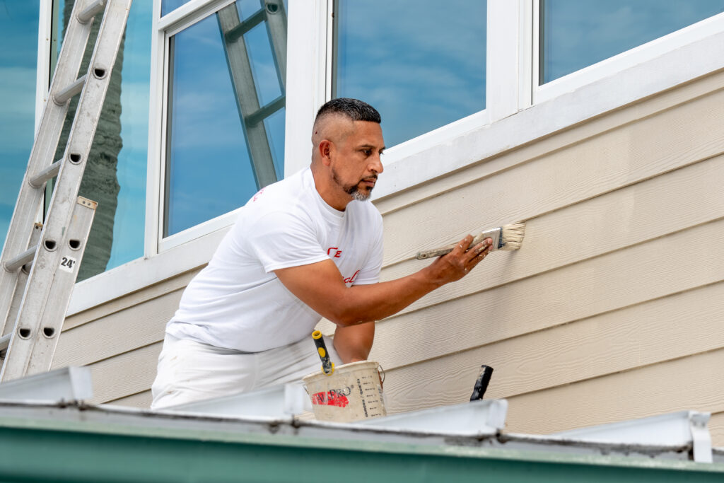 Professional painter in white brushing exterior siding beside white-trimmed windows, with a ladder and paint bucket nearby, while painting in Florida humidity